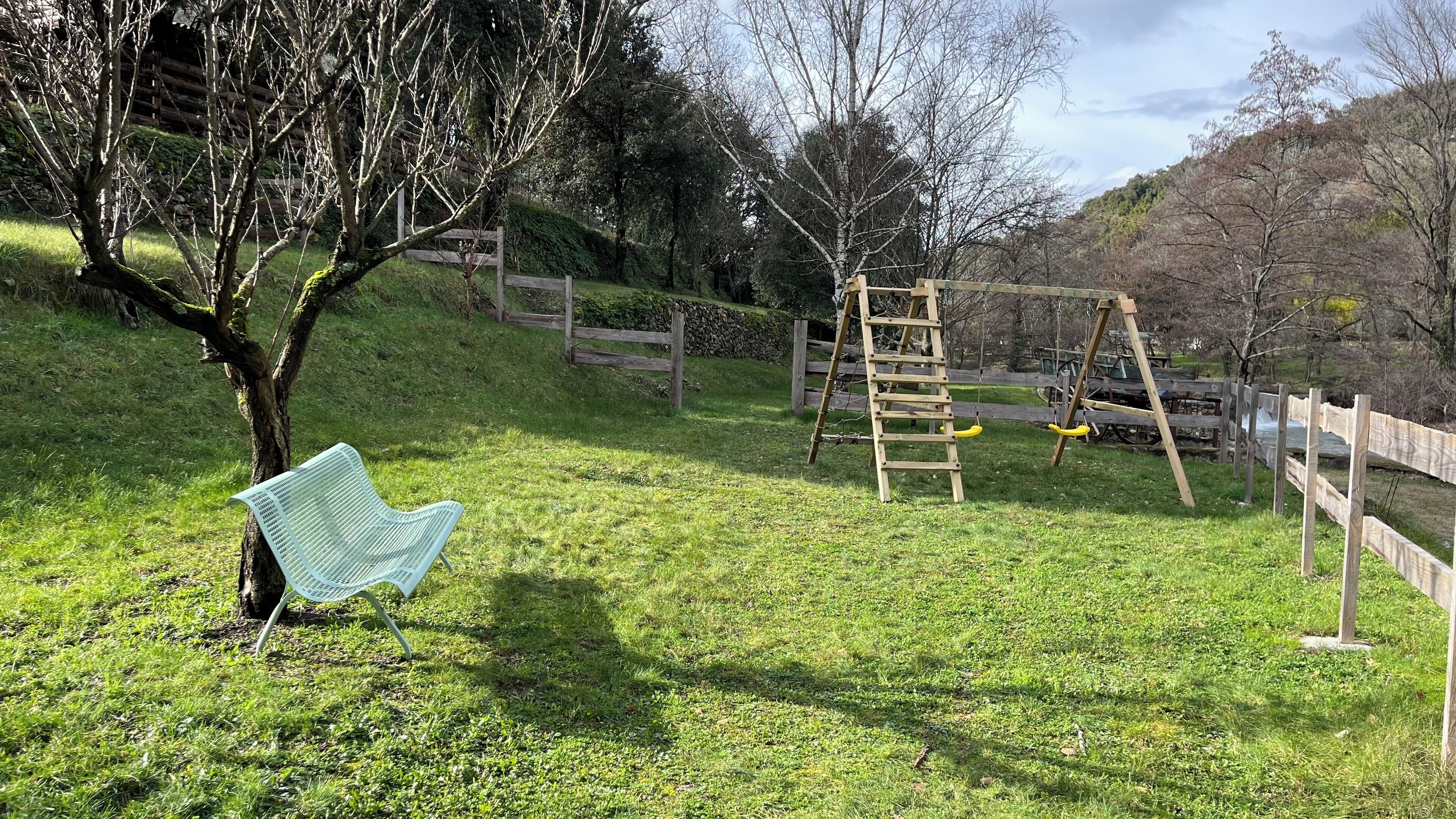Un espacio al aire libre con césped, un columpio de madera, un banco de metal y un árbol.