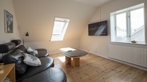 Un salon moderne avec un canapé en cuir noir, une table basse en bois, un lampadaire et une grande fenêtre donnant sur des immeubles.
