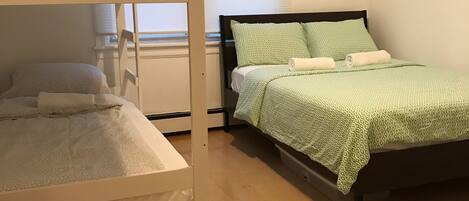 A room with two bunk beds, one with a white frame and the other with a dark frame. There is a window with blinds, a radiator, and a wooden floor.