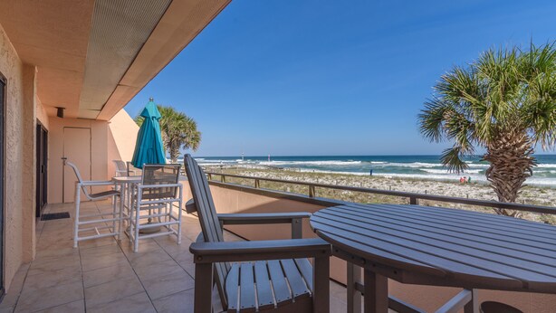 A balcony with a table and chairs, overlooking a beach with palm trees and the ocean.