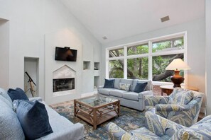 A living room with a fireplace, a television, and a large window with a view of trees. - Direct Beach Path & Pool Near The Salty Dog Cafe (Hilton Head Island)