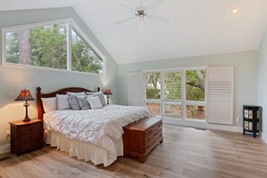 A bedroom with a large bed, two bedside tables with lamps, a dresser, a ceiling fan, and a large window with shutters.