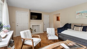 A modern bedroom with a sofa bed, two white armchairs, a flat-screen TV, a fireplace, and a wooden coffee table.