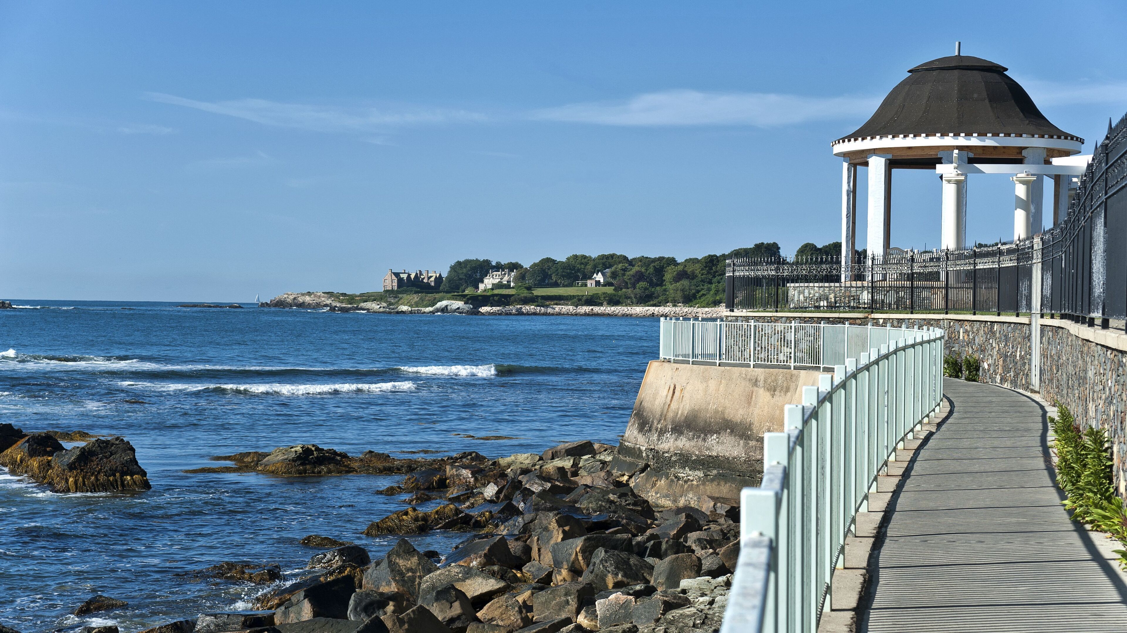Un paseo marítimo con un quiosco, costa rocosa y vistas de casas a lo lejos.