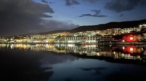A waterfront town with buildings reflecting on calm water, a mountain in the background, and a red-lit structure on the right.