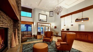 A lobby with a stone wall, a fireplace, leather armchairs, a round ottoman, a chandelier, and a reception desk with a sign. - Stoneridge 2 BDR - Sleeps 8★ Hot Tub/ Pool/Sauna/Gym ★Mountain Views ★1100 SQF (Canmore)
