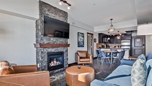 A modern living room with a stone fireplace, leather furniture, and a flat-screen TV mounted on the wall. - Stoneridge 2 BDR - Sleeps 8★ Hot Tub/ Pool/Sauna/Gym ★Mountain Views ★1100 SQF (Canmore)