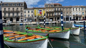 Un port de plaisance où sont amarrés plusieurs petits bateaux, une rangée de bâtiments abritant divers commerces et affichant différentes enseignes, et un ciel bleu dégagé.