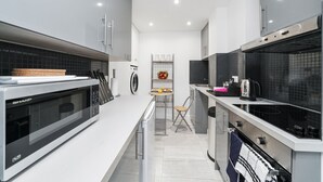 A modern kitchen with a microwave, toaster, and kettle. There is a washing machine and a small dining area with a chair and table. - Cosy Apartment in the Heart of the City (London)
