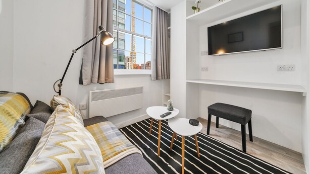A modern bedroom with a bed, bedside lamp, window with curtains, radiator, small round table, bench, and a TV on the wall. - Cosy Apartment in the Heart of the City (London)