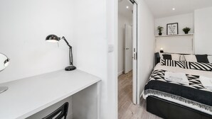 A modern bedroom with a white desk, a black desk lamp, a bed with black and white striped bedding, and a small shelf with decorative items. - Cosy Apartment in the Heart of the City (London)