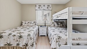 A bedroom with two single beds, a bunk bed, a nightstand, a lamp, and a window with blinds. - Ocean Garden Villas M2-2BR/2BA Oceanfront Condo in Crescent Beach (North Myrtle Beach)
