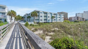 A wooden boardwalk leading to a beachfront building with multiple balconies and a beach in the background. - Ocean Garden Villas M2-2BR/2BA Oceanfront Condo in Crescent Beach (North Myrtle Beach)