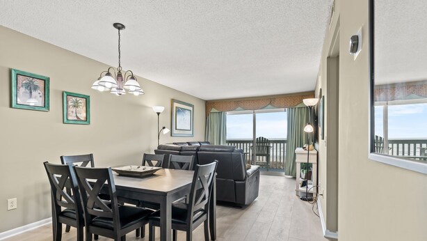 A modern living room with a dining table, chairs, a sofa, and a balcony with a view. - Ocean Garden Villas M2-2BR/2BA Oceanfront Condo in Crescent Beach (North Myrtle Beach)