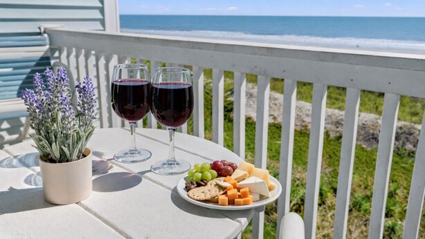 Una mesa con una bandeja de quesos, dos copas de vino tinto y una maceta de lavanda en un balcón con vistas al océano.