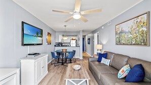 A modern living room with a flat-screen TV, a ceiling fan, a dining area with blue chairs, and a kitchen in the background. - Ocean Pier I 107-2BR/2BA Oceanfront Condo in Windy Hill (North Myrtle Beach)