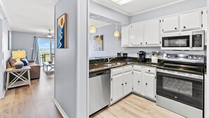 A modern kitchen with white cabinets, a black stove, and a microwave. There is a dining area with a table and chairs, and a view of the outdoors through a window. - Ocean Pier I 107-2BR/2BA Oceanfront Condo in Windy Hill (North Myrtle Beach)
