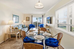 A dining area with a glass table set for four, surrounded by wicker chairs with blue cushions, a sofa with floral upholstery, and a television on the wall. - Eat, Beach, Sleep and Repeat at Ocean Pier III 128 (North Myrtle Beach)