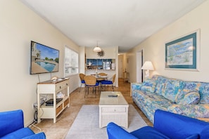 A living room with a sofa, a dining table with chairs, a kitchen area, and a painting on the wall. - Eat, Beach, Sleep and Repeat at Ocean Pier III 128 (North Myrtle Beach)