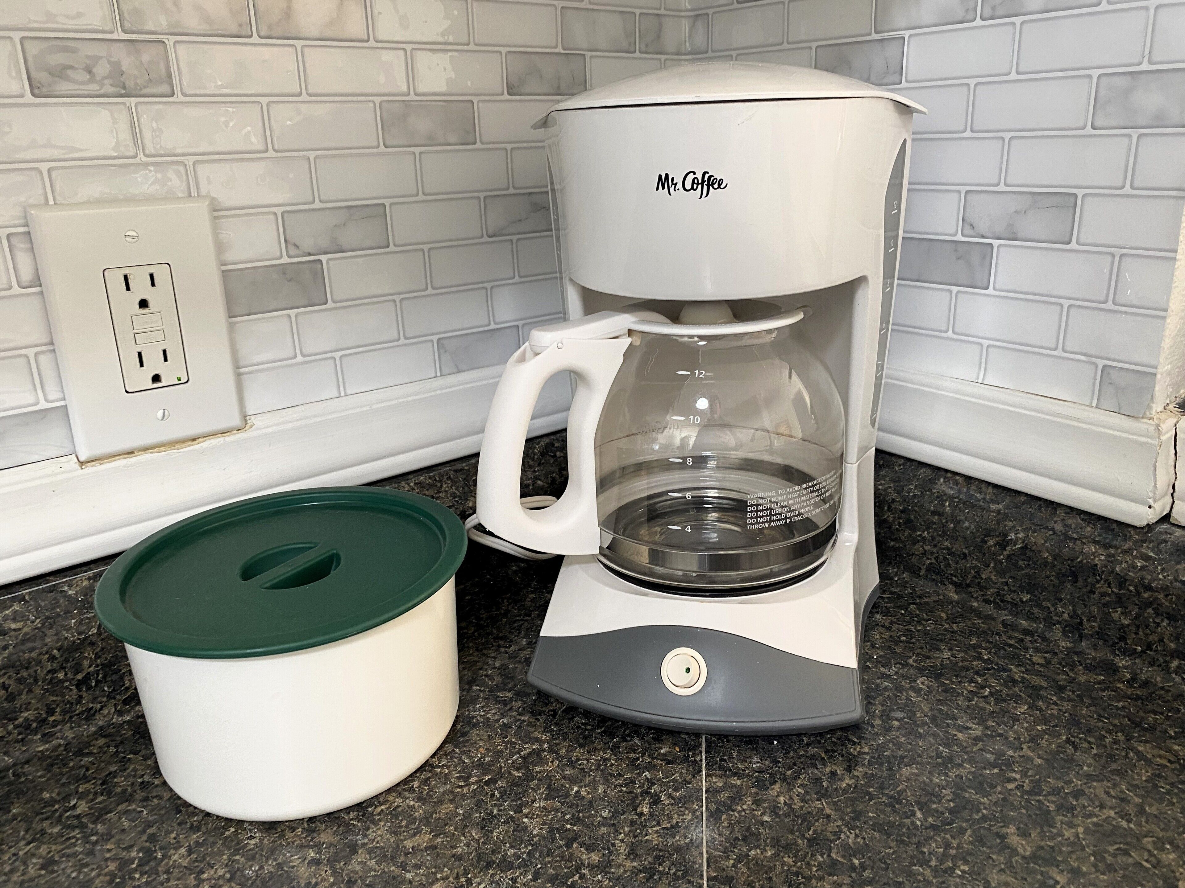A coffee maker with a green lid and a clear carafe, placed on a granite countertop next to a white electrical outlet.