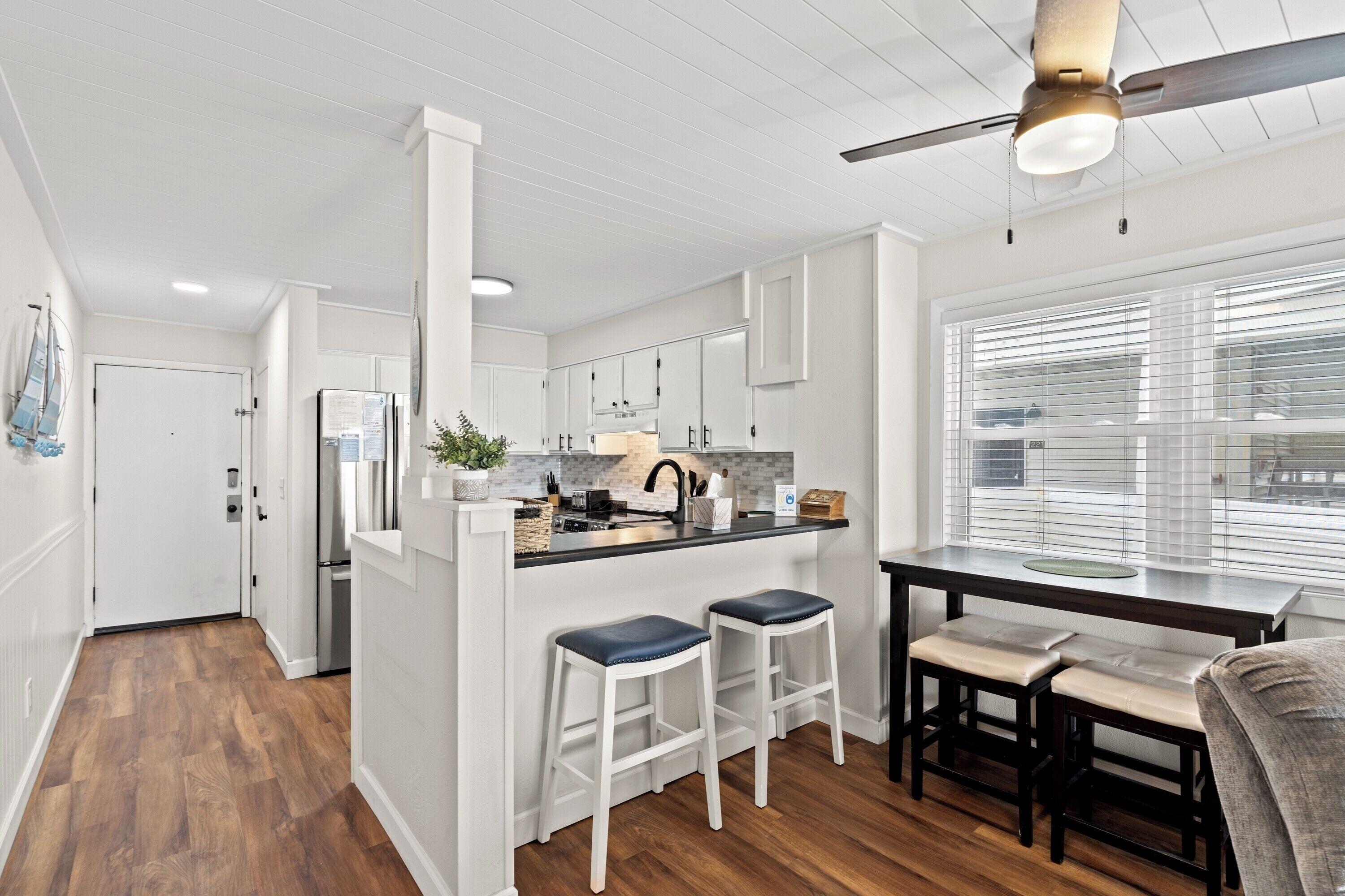 A modern kitchen with white cabinetry, a black countertop, and a ceiling fan. There are two bar stools and a dining table with chairs.