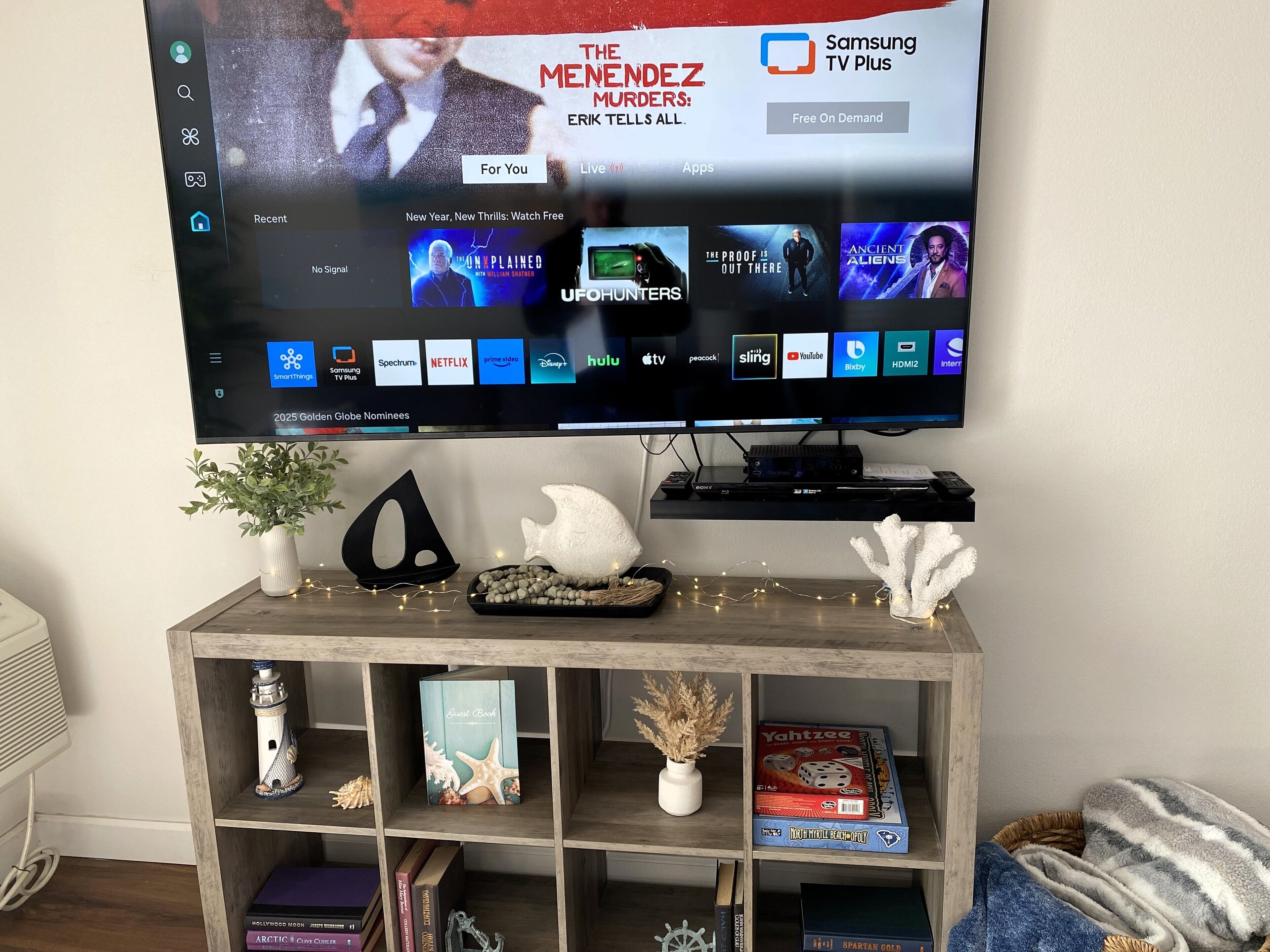 A flat-screen TV mounted on a wall displaying a streaming service interface with various movie options. Below the TV, there is a wooden shelf with decorative items and books.