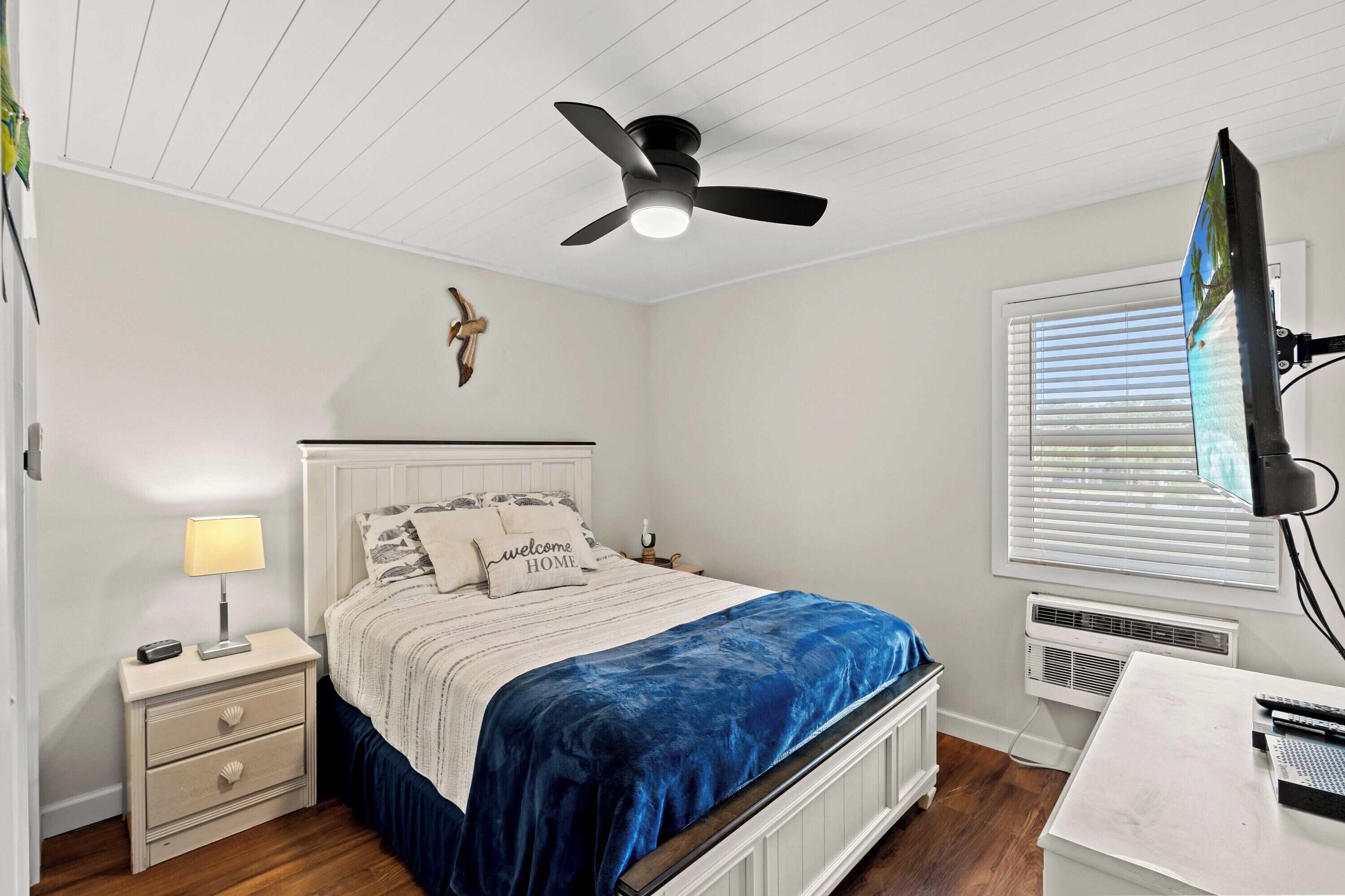 A bedroom with a bed, nightstand, lamp, ceiling fan, TV, and window with blinds.