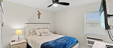 A bedroom with a bed, nightstand, lamp, ceiling fan, TV, and window with blinds.