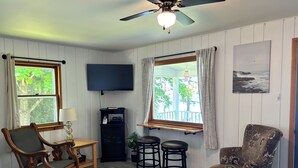 A living room with a TV, wooden furniture, a ceiling fan, and a window with a view of trees.