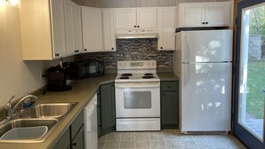A compact kitchen with white cabinets, a white stove, a white refrigerator, a stone backsplash, and a window with a view of the outdoors.