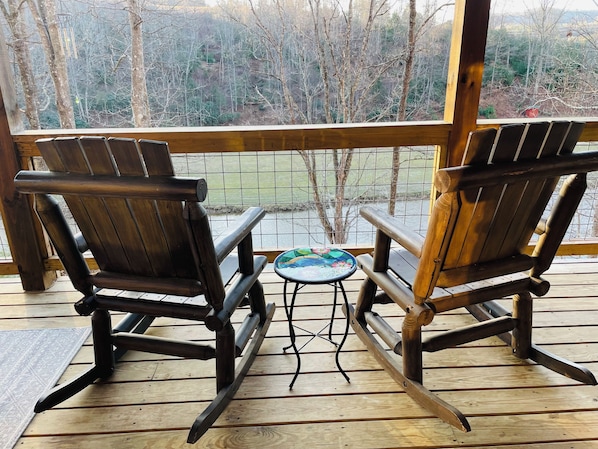 Dos sillas de madera tipo Adirondack mirando hacia una mesa pequeña con mantel floral en una terraza de madera con vistas a un área boscosa.