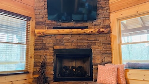 Ein gemütliches Zimmer mit einem Steinkamin, einem Wand-Fernseher und einem Fenster mit Jalousien.