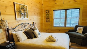 Ein gemütliches Holzblockhaus-Zimmer mit Bett, Nachttisch, Kommode, einem Fenster mit Jalousien und einer Couch.