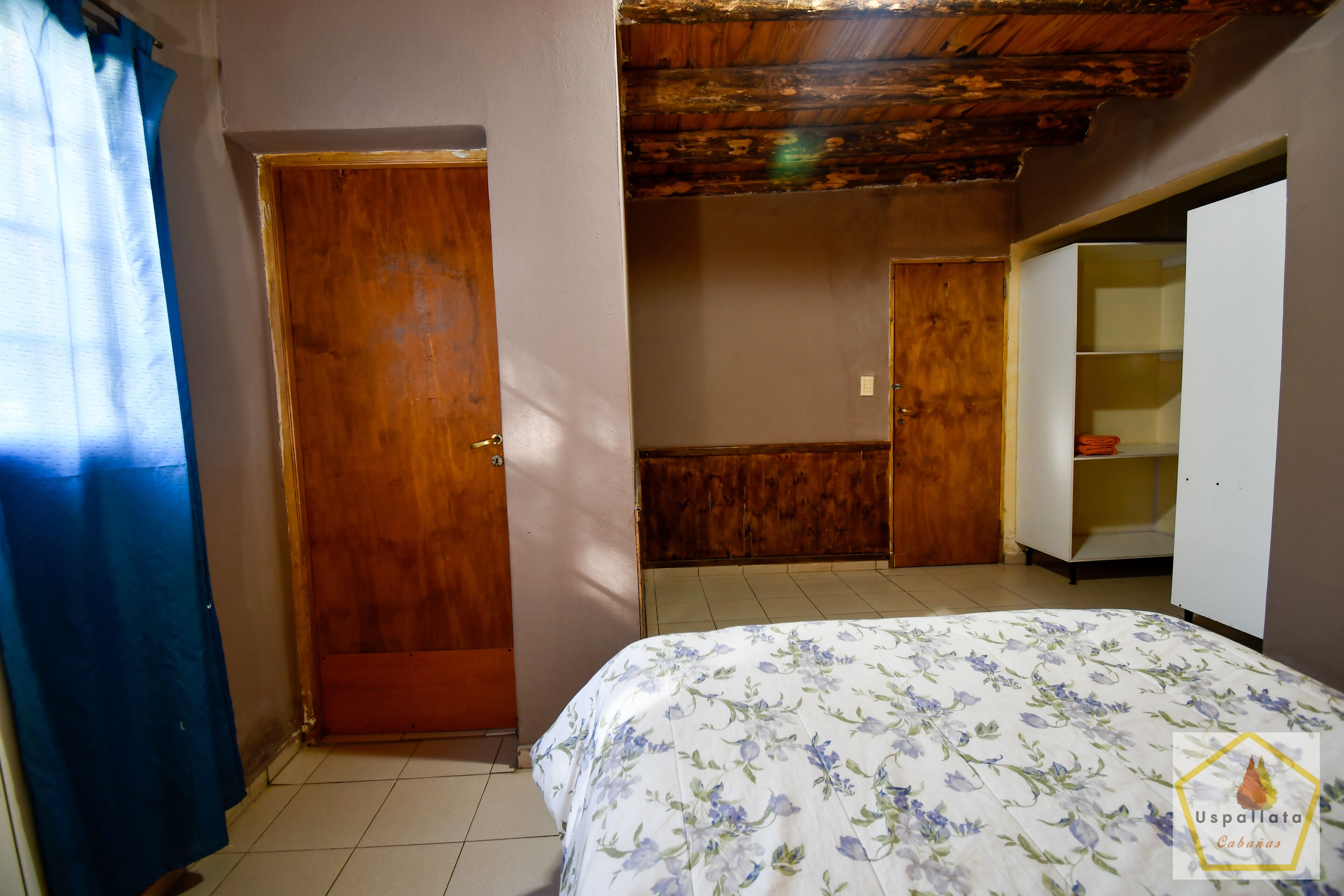 Une chambre avec une porte en bois, un lit recouvert d’une couvre-lit à motifs floraux et une armoire.