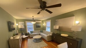 Una sala de estar con sofá modular, una mesa de centro, televisor, ventilador de techo y chimenea.