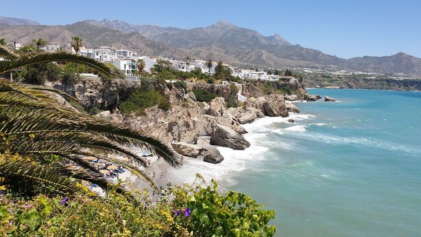 A coastal town with rocky cliffs, a beach with umbrellas, and a mountain range in the background.