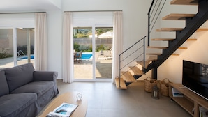 Una sala de estar moderna con un sofá gris, una mesa de centro de madera, un televisor y una escalera que lleva a un área exterior con piscina.