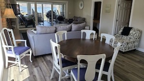Ein großzügiger Wohnbereich mit einem runden Holztisch, umgeben von weißen Stühlen mit blauen Kissen, einem grauen Sofa und einem gemusterten Sessel.