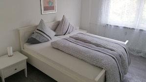 Un dormitorio ordenado con cama, almohadas, mesita de noche, un cuadro enmarcado en la pared y una ventana con cortinas translúcidas.