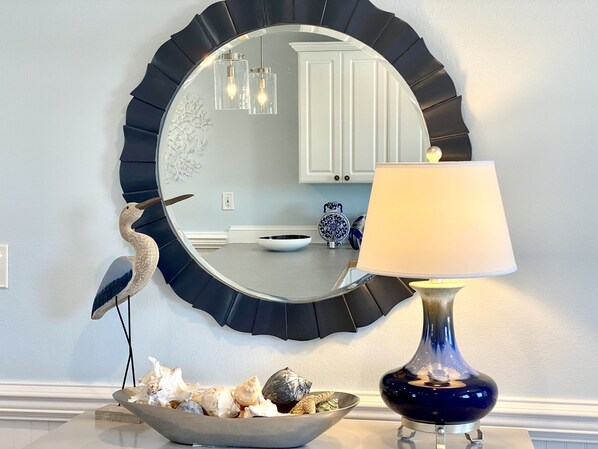 A round mirror with a black frame, a table with a bowl of seashells, and a blue lamp.