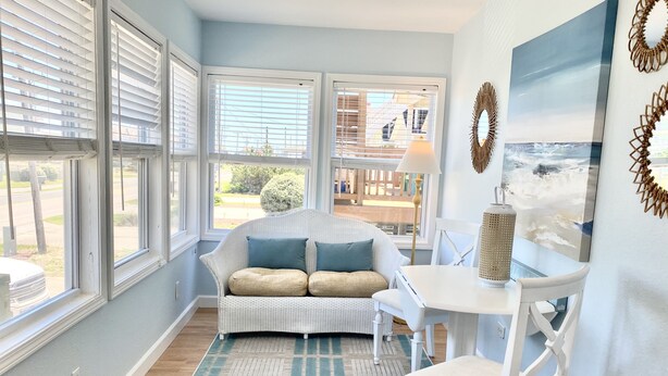 A room with a white wicker sofa, a white table, and a chair, a patterned rug, and a view through large windows.