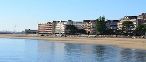 A beachfront hotel with multiple floors, a sandy beach in the foreground, and a clear sky.