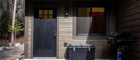A wooden house with a black door, a window, and a barbecue grill on the porch.