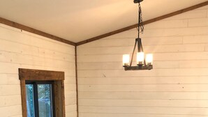 Een kamer met een ronde houten tafel, twee krukken, een raam met houten kader en een hangende lamp.