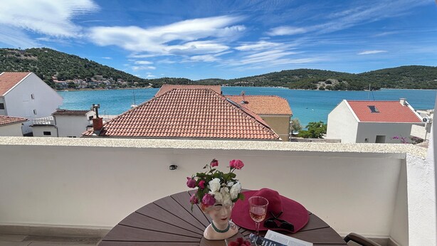 Un balcón con vistas a un pueblo costero, una mesa con un jarrón de flores, una copa de bebida y un libro.