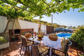 An outdoor patio with a glass table, wicker chairs, a stone fireplace, and a swimming pool in the background.