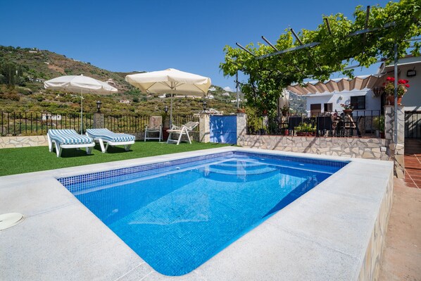A rectangular swimming pool with blue tiles, surrounded by a stone wall. There are two lounge chairs with striped cushions and a white umbrella nearby.