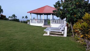 A gazebo with a red roof and white columns, surrounded by a well-maintained lawn and tropical plants. - Villa en Alquiler con Vista al mar (Cabrera)