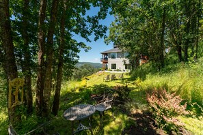 Une maison dotée d’une terrasse, entourée d’arbres et bordée d’une pelouse où se trouvent deux tables et des chaises en métal.