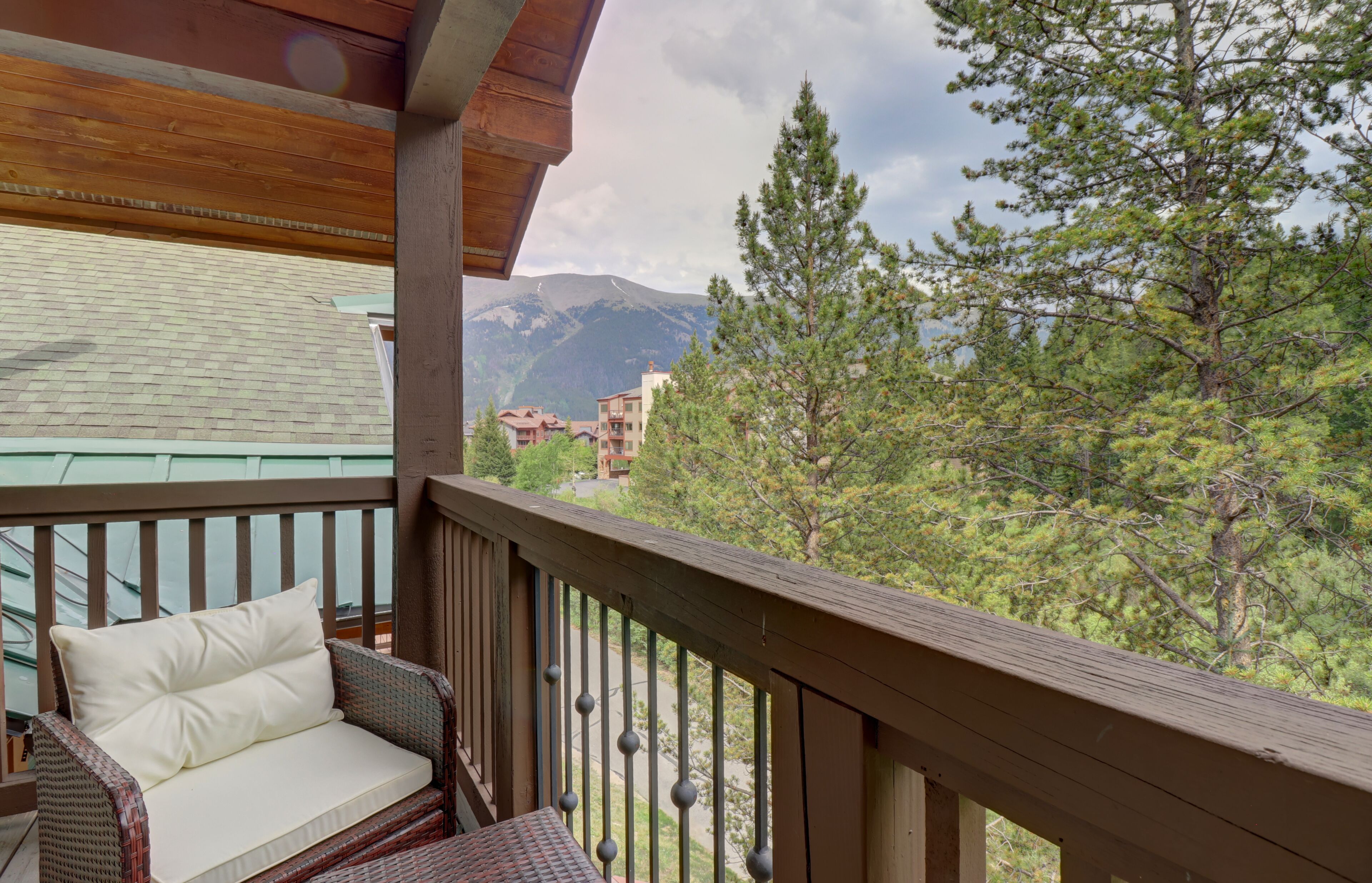 A balcony with a wicker chair and a cushion, overlooking a mountainous landscape with trees and buildings.